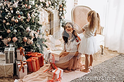 Happy young mother and her little daughter in nice dress sit near the New Year`s tree and open New Year`s gifts in the Stock Photo