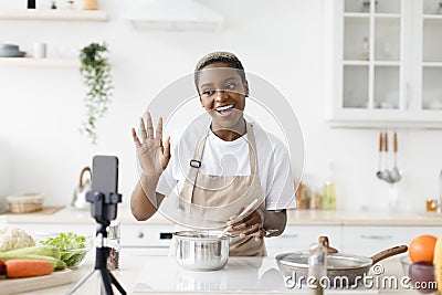 Happy young pretty black woman blogger in apron prepares dinner, shows dish, waves hand, shoots video for blog Stock Photo