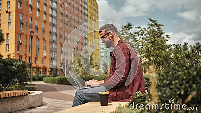 Happy and young. Side view of handsome man with stubble in casual clothes and eyeglasses working on laptop while sitting Stock Photo