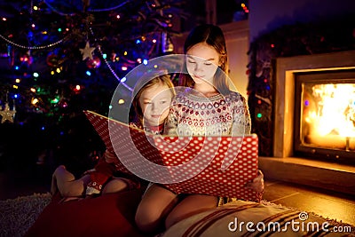 Happy young sisters reading a story book together by a fireplace in a cozy dark living room on Christmas eve. Celebrating Xmas at Stock Photo