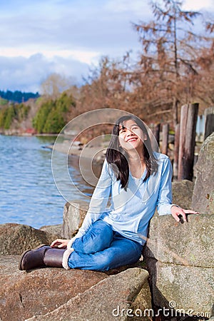Happy young teen girl face upturned, smiling, while sitting outdoors on rocks along lake shore Stock Photo