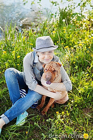 Happy young woman in denim overalls and hat hugging his beloved dog Shar Pei in the green grass in sunny day, true friends forever Stock Photo