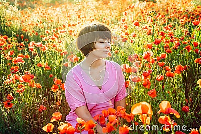 A happy young woman in a pink dress with closed eyes, relaxing in red poppies flowers meadow in sunset light. A simple pleasure Stock Photo