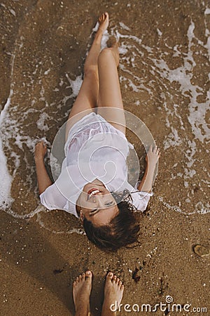 Happy young woman in wet white shirt lying on beach in splashing waves. Top view. Stylish tanned girl relaxing on seashore and Stock Photo