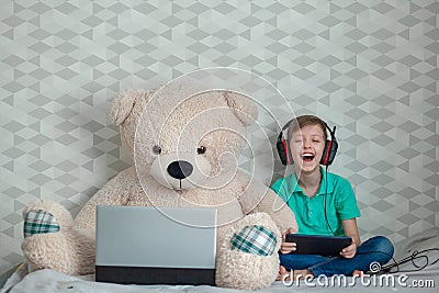 Happykid in headphones playing on digital tablet next to a toy bear looking at a computer Stock Photo