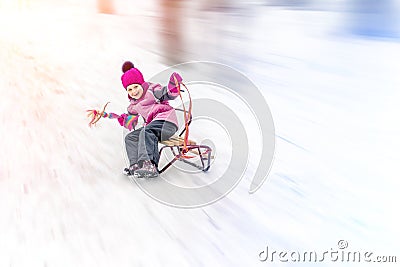 Hapy cutee litlle girl having fun sledging from hill on bright sunny day. Child moving fast on sledges downhill at Stock Photo