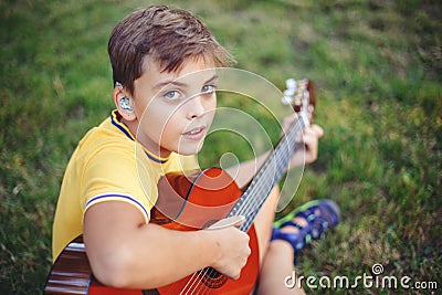 Hard of hearing preteen boy playing guitar outdoors. Child with hearing aids in ears playing music and singing song in park. Hobby Stock Photo