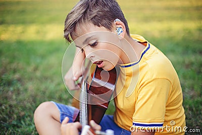 Hard of hearing preteen boy playing guitar and singing. Child with hearing aids in ears playing music and singing song in a park. Stock Photo