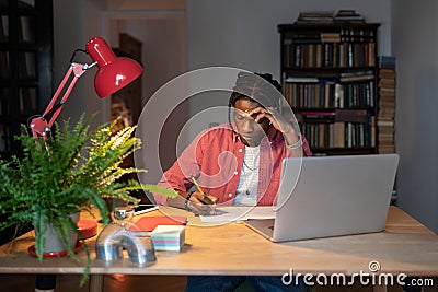 Hardworking young African guy student writing notes while watching webinar on laptop at home Stock Photo