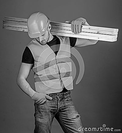 Hardy labourer concept. Man in helmet, hard hat and protective gloves holds wooden beam, grey background. Carpenter Stock Photo