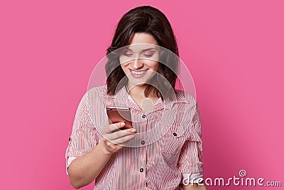 Ð¡harming, beloved female person looks into smartphone with smile in middle of portrait photography on isolated pink background. Stock Photo