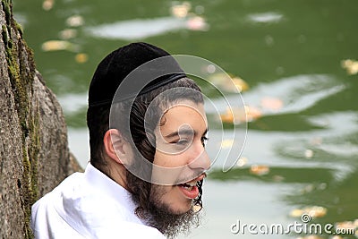 The Hasid boy in a traditional pile sits near a lake in the park. Editorial Stock Photo