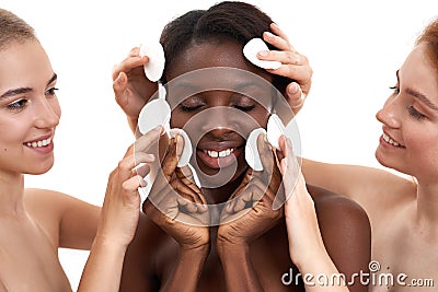 Having fun together. Two young and happy caucasian women help to clean face of their african female friend with cotton Stock Photo