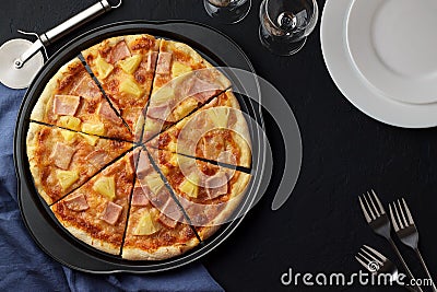 Hawaiian pizza with sweet pineapple and salty ham on dark stone background with plates, forks and glasses. Stock Photo