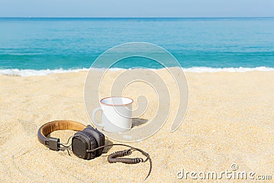 Headphones and coffee mugs placed on the sand with sea and sky background. Travel concept with copy space. Stock Photo