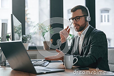 In headphones. Having online conversation. Businessman in suit is in the office with computer Stock Photo