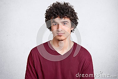 Headshot of attractive curly youngster looks seriously at camera, has appealing look, spends free time with friends, dressed in ca Stock Photo