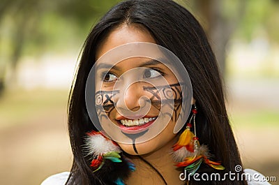 Headshot beautiful Amazonian woman, indigenous facial paint and earrings with colorful feathers, posing happily for Stock Photo