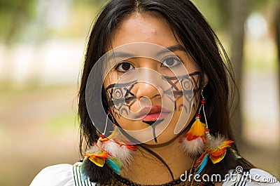 Headshot beautiful Amazonian woman, indigenous facial paint and earrings with colorful feathers, posing seriously for Stock Photo