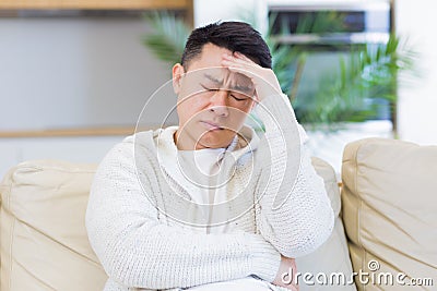 Asian man holding his head with a severe headache at home in a room on the couch. Up close, the casual male Stock Photo