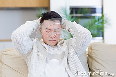 Asian man holding his head with a severe headache at home in a room on the couch. Up close, the casual male Stock Photo