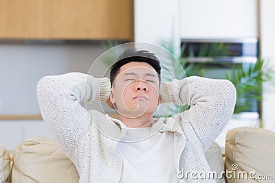 asian man holding his head with a severe headache at home in a room on the couch. Up close, the casual male Stock Photo