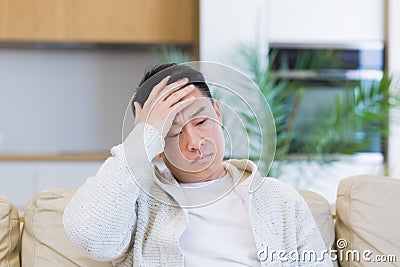 asian man holding his head with a severe headache at home in a room on the couch. Up close, the casual male Stock Photo