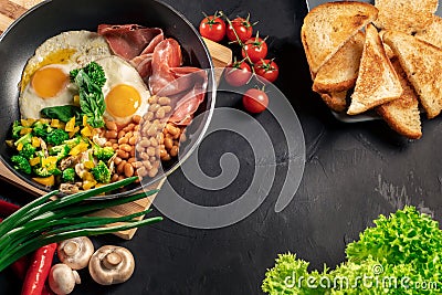 Healthy breakfast with fried eggs, bacon, beans, toasts, mushrooms, broccoli and fresh salad on black background Stock Photo