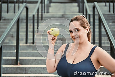 Healthy life. Positive plus size woman in sport clothing holding green apple and smiling while standing on stairs Stock Photo