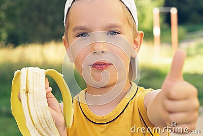 Healthy little boy eating banana. Happy kid enjoy eating fresh fruit and showing thumbs up. Stock Photo