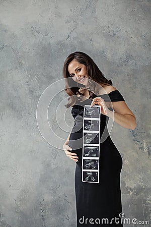 Healthy pregnant woman holding ultrasound scan image, smiling and embracing her tummy on gray Stock Photo