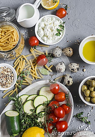 Healthy vegetarian food ingredients - pasta, zucchini, tomatoes, herbs, spices, vegan cheese, olive oil, lemon, olives. On a gray Stock Photo