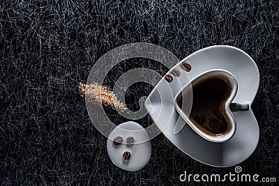 A heart shaped coffee cup with coffee beans, spilled milk and brown sugar on a black background with silver lining Stock Photo