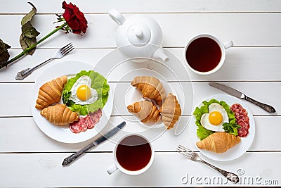 Heart shaped fried eggs, salad, croissants, salami sausage, rose flower composition and tea, white wooden table Stock Photo