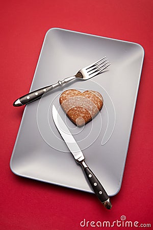 Heartbreaker concept image: heart shaped cookie on plate with fork and knife over red background Stock Photo