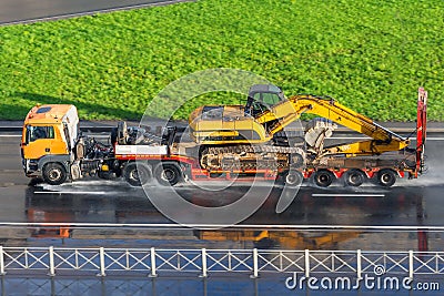 Heavy yellow excavator on transportation truck with long trailer platform on the highway wet asphalt after rain in the city Stock Photo