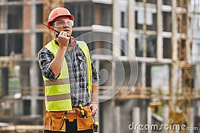 Hecking work. Professional young builder in working uniform and red protective helmet talking to crane operator by Stock Photo