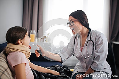 Helpful young emale doctor sit beside patient in room. She hold spay for soring throat. Doctor show to girl how keep Stock Photo