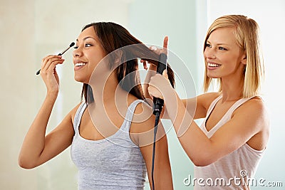 Helping her to look her best. a young woman applying makeup while her friend straightens her hair. Stock Photo