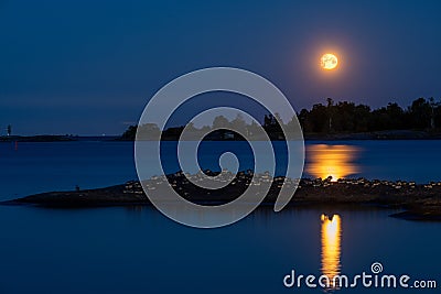 A beautiful full moon casting reflection on the calm water. A silhouette of a small skerry with a flock of Canada Gooses Stock Photo