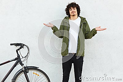 Hesitant curly man shruggs shoulders in bewilderment, dressed in fashionable green anorak, stands near bicycle, isolated over Stock Photo