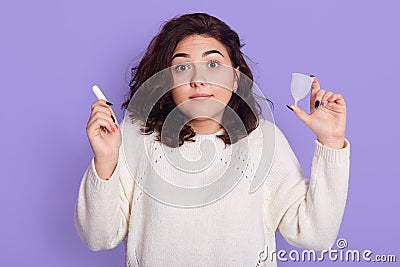 Hesitant dark haired woman thinks what better to choose during critical days, holds menstrual cup and tampon, looks astonished at Stock Photo