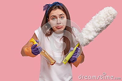 Hesitant displeased woman holds duster and cleaning liquid or spray, feels apathy, doesnt want to tidy up room, wears Stock Photo