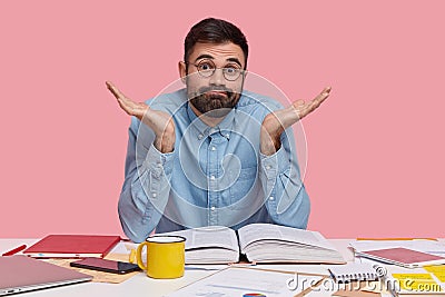 Hesitant unshaven man shruggs shoulders, has clueless facial expression, dressed in formal wear, round spectacles Stock Photo