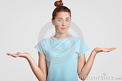 Hesitant young woman shrugs shoulders with uncertainy, cant make decision, clasps palms, looks clueless at camera, dressed in blue Stock Photo