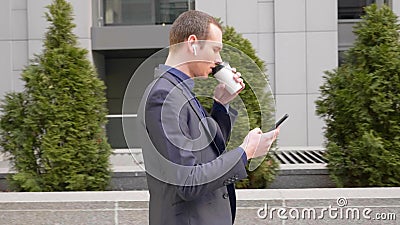 Het jonge zakenman lopen onderaan de straat met draadloze oortelefoons in de oren en schrijft een bericht op de telefoon stock footage Het jonge zakenman lopen onderaan de straat met draadloze oortelefoons in de oren en schrijft een bericht op de telefoon stock footage
