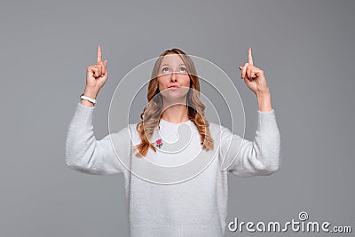 Hey look there. Beautiful blonde young woman pointing fingers up, looking amazed, showing something interesting, stands in white Stock Photo