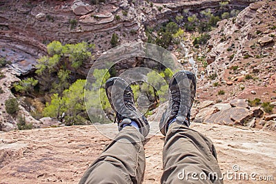 A hiker dangles his feet over the edge of a cliff looking down into a desert canyon. Stock Photo