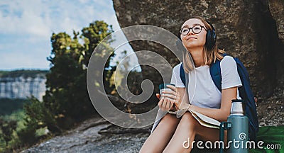 Hiker girl with backpack and Ñup of drink in her hand sitting in mountains and plans trip route on map, female person in glasses Stock Photo