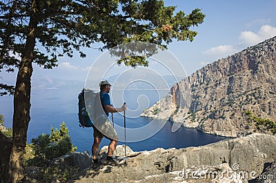 Hiking on Lycian way. Man with backpack enjoys view of Butterfly Valley blue lagoon from rocky cliff in shadow Stock Photo